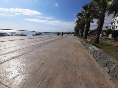 The image shows a promenade in Atakum, Turkey, on a sunny day with a bright blue sky and scattered clouds. The promenade is paved with large, textured concrete tiles and runs alongside a calm body of water, likely the Black Sea, where several boats are docked. Palm trees line the right side of the promenade, separating it from a grassy area and a street with buildings. People can be seen walking and jogging on the promenade. In the distance, a cityscape is visible. The scene conveys a sense of a pleasant, active outdoor environment.