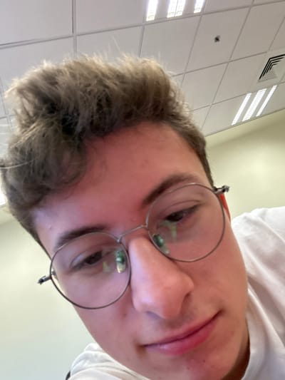 A close-up selfie shows a young person wearing round glasses and a white hoodie. The camera angle is low, looking up at the person's face. The background is a brightly lit ceiling with white square tiles and recessed lighting. The person's expression is neutral with a slight smile. Their hair is short and light brown. The glasses reflect the ceiling lights.