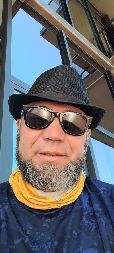 A man wearing a black fedora, black sunglasses, and a blue patterned shirt with a yellow neck gaiter is looking at the camera. He has a grey beard and is outdoors with a blue sky and a building in the background. The image appears to be a selfie taken at an outdoor location, possibly a restaurant or balcony in Milisi, Georgia, given the architectural style of the building and the clear sky.