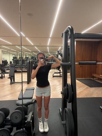 A woman in a black t-shirt and gray shorts poses, flexing her arm in a gym mirror. Her phone is visible in her hand. Dumbbells and weight machines fill the background. The gym has a wood floor and light-colored walls. A weight machine is visible above her.