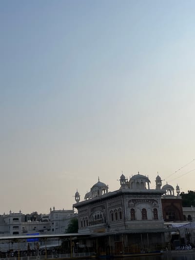The image depicts the Golden Temple, also known as Sri Harmandir Sahib, in Amritsar, India. The foreground shows the reflective waters of the Amrit Sarovar, a sacred pool surrounding the temple complex. A walkway with people is visible in the lower section of the image, suggesting a public gathering. The temple itself, characterized by its white marble facade and ornate architectural details, rises majestically from the water. Several domes and minarets punctuate the skyline. The background reveals a serene sky with a gradient of light blue to pale yellow, indicating either early morning or late afternoon. The overall atmosphere is peaceful and spiritual, befitting a holy site.