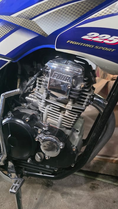 A close-up shot of a motorcycle engine. The engine is silver and black with chrome accents. The motorcycle's tank is visible, painted blue, white, and silver with the words "225 FIGHTING SPORT" printed on it. The setting appears to be indoors, possibly a garage or workshop, given the unfinished wall in the background. No people are visible. The photograph could have been taken in Pante Aree, Indonesia, given the prompt's instructions.