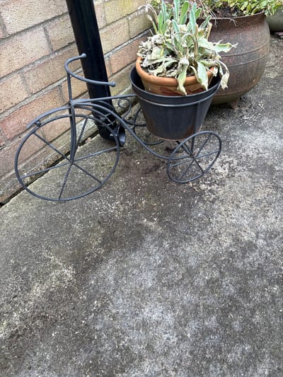 The image shows an outdoor scene, possibly in Longden, United Kingdom, given the prompt's location reference. The foreground is comprised of a brick surface on the lower left and a gray concrete slab dominating the rest of the scene. A black metal plant stand, shaped like a bicycle, is partially visible, holding a brown terracotta pot within a black metal container. The plant inside the pot appears to be dried or withered. To the upper left, a large, rusted metal pot rests on the brick surface, partly obscured by the plant stand. The concrete slab exhibits a weathered texture with visible cracks and stains.