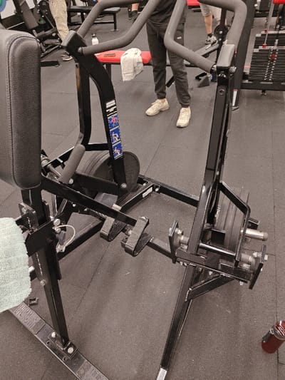 An indoor, close-up shot depicts a black seated calf raise machine at a gym. The machine features pads for foot placement, weight plates on rotating bars, and a padded seat. A light green towel rests on the edge of the seat. In the background, gym patrons and other exercise equipment can be seen. The floor is dark and made of rubber. A red water bottle is visible on the bottom right.