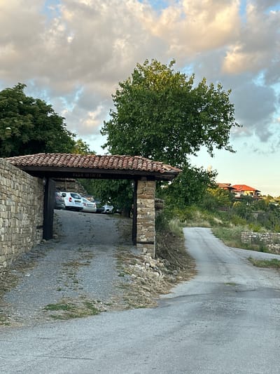 This image depicts a stone archway entrance to a property in Arbanassi, Bulgaria. The archway is constructed with rough-hewn stones and topped with a tiled roof. A wooden beam above the entrance is inscribed with Cyrillic text, "КЪЩА ЗА ГОСТИ," which translates to "Guest House."
Inside the archway, a gravel driveway leads to several parked vehicles, including a white van and two cars. To the right of the archway, a paved road curves uphill and disappears into the distance. The surrounding environment is characterized by lush green trees, dense vegetation, and more stone walls. In the background, houses with red-tiled roofs are visible on a hillside, suggesting a residential area. The sky above is partly cloudy, with the clouds illuminated by warm, soft light, indicating either early morning or late afternoon. The overall impression is one of a rustic and traditional Bulgarian setting.