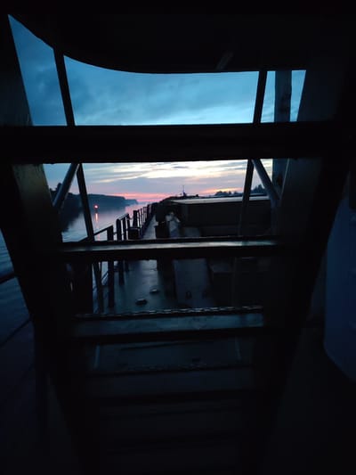 The image depicts a view from within a dark, possibly industrial structure, looking out at a tranquil body of water during dusk. The foreground is dominated by dark, silhouetted metal beams and railings, suggesting the perspective is from inside a ship, a bridge, or a similar edifice.
Through the framing elements, a serene river or lake landscape is visible. The water is calm, reflecting the soft hues of the twilight sky. On the opposite bank, a treeline is discernible, with a distant landmass extending into the distance. A small red light, possibly a buoy or a distant structure, is visible on the water.
The sky exhibits a gradient of colors characteristic of sunset or sunrise, with streaks of pink, orange, and blue clouds against a darkening blue canvas. The overall atmosphere is peaceful and contemplative, capturing a moment of transition between day and night. The location specified, Badozhskoi Pogost, Russia, places this scene within a rural or semi-rural setting, likely characterized by waterways and natural landscapes.