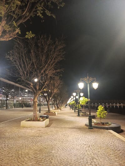 This image captures a night scene on a cobblestone pathway in Al Machouar – Stinia, Morocco. The pathway is lined with trees planted in concrete or stone planters, and classic lampposts illuminate the area. The trees near the camera are bare-branched, while some further down the path have foliage. In the background, a body of water reflects distant lights, and a building with a modern facade is partially visible on the left. Several people are walking in the distance. The sky is dark, suggesting a late hour.