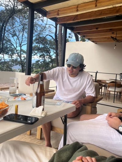 An outdoor scene features a man in a white t-shirt and light blue cap, wearing sunglasses, at a table with a beer bottle and snacks. Trees and a building structure are visible in the background. The scene includes other people seated, one wearing a watch, suggesting a relaxed social gathering. A mobile phone also sits on the table.