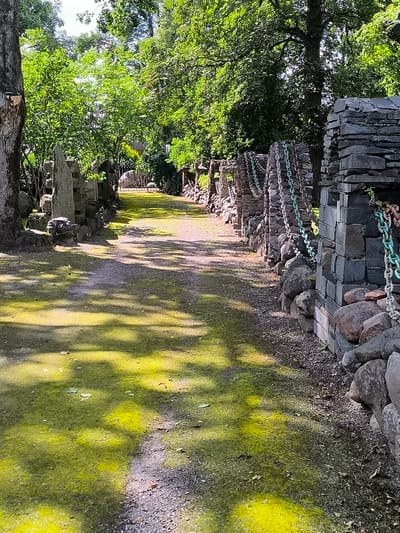The image depicts a pathway leading through a park-like setting in the city of Mosėdis, Lithuania. The path, composed of gravel and patches of bright green moss, is flanked by stone structures. On the right side of the path, a wall constructed from stacked flat stones rises, adorned with several lengths of thick metal chain, some green and some silver. Large, rounded stones are integrated into the base of this wall. On the left side, the pathway is bordered by a similar arrangement of stone pillars and rocks, with some larger, more irregularly shaped standing stones visible. Trees with dense green foliage form a canopy overhead, casting dappled shadows onto the path. In the distance, a wrought-iron gate can be seen, suggesting an entrance to another area. The overall impression is one of a rustic, natural, and somewhat artful outdoor space.
