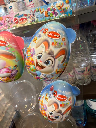 In Ravno pole, Bulgaria, a close-up shot reveals an Easter-themed display, likely in a retail setting, featuring two "Mad Max Lucky Egg" Easter eggs. The egg in the foreground, with a blue background, showcases a cartoon rabbit character holding colorful Easter eggs, alongside text that reads "Happy Easter." Adjacent to it, a pink Easter egg with a similar cartoon theme is partially visible on the left. Behind the eggs, clear plastic packaging and several bottles of water are present, suggesting a product display on a shelf. The lighting is bright, highlighting the vibrant colors of the Easter egg designs.