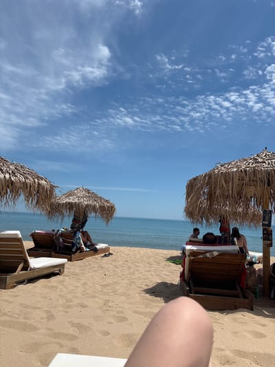 The image depicts a sunny day at the beach in Shkorpilovtsi, Bulgaria. The scene is oriented vertically, with the sky occupying the left two-thirds and the beach, sea, and seating area on the right. The sky is a vibrant blue with scattered white clouds, suggesting fair weather.
In the foreground, on the sandy beach, are several thatched umbrellas providing shade over wooden beach chairs. Some people are visible lounging on these chairs. In the mid-ground, the calm, blue sea stretches to the horizon, meeting the sky. The sand is light brown and appears undisturbed except for some footprints.
On the right side of the image, a person's leg is visible in the very foreground, suggesting the photo was taken from the perspective of someone lying down. Several wooden beach loungers are arranged under thatched umbrellas, with people seated or lying on them. One lounger has a wooden structure, possibly for storage. Colorful towels and bags are placed on and around the loungers.
The time of day appears to be daytime, given the bright sunlight and clear sky. The atmosphere seems relaxed and leisurely, typical of a beach vacation. No specific events are taking place beyond people enjoying a day at the beach.
There are some signs visible on a wooden post near the top of the image, but the text is too small and blurry to be legible. The location is identified as Shkorpilovtsi, Bulgaria.