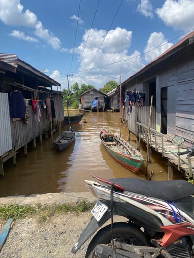 The image depicts a waterfront scene in Berangas Timur, Indonesia, likely a village on the coast or a river. Two rows of wooden stilted houses line a narrow waterway. Laundry hangs on lines strung between houses on the left. Several small wooden boats are moored in the brown water, some close to the houses and others further out. One larger boat with red and green trim is prominent in the middle ground. The sky is blue with scattered white clouds. A portion of a motorcycle, with a license plate visible that reads "6922 LY 08-27", is in the foreground. The overall impression is of a traditional, water-based community.