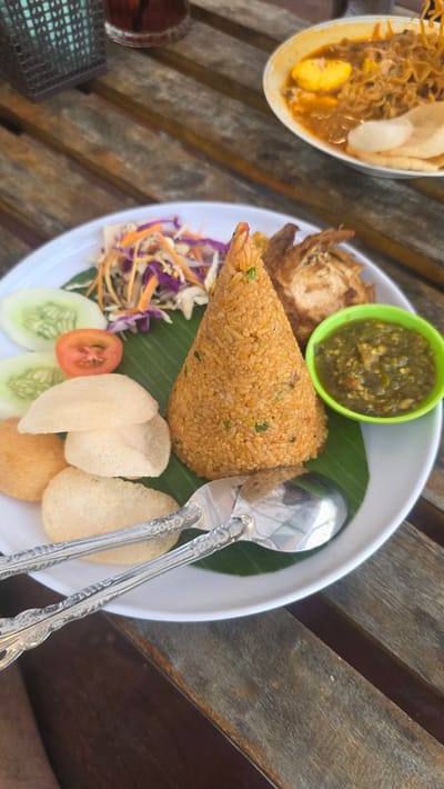 The image depicts a meal served on a white plate, presented on a wooden table. The primary element of the meal is a conical mound of orange fried rice. Accompanying the rice are several side items: a small green bowl of chutney or relish, a fried egg or omelet, a shredded salad of purple and orange cabbage, slices of cucumber and tomato, and several prawn crackers. A pair of silver spoons are laid across the plate. To the left of the main plate, there is another dish containing noodles and what appears to be a hard-boiled egg. A small glass of liquid is also visible. The image was taken in the city of Kuala Pidie, Indonesia.
