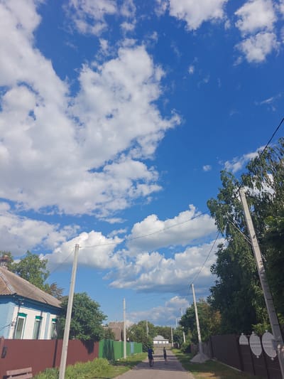 The image depicts a street scene in Kotelva, Ukraine, under a bright blue sky with scattered white clouds. On the left, a two-story house with a blue and white facade and a grey roof is visible. A brown fence runs along the front of the property. A rural road stretches into the distance, lined with trees and fences. Two individuals are present on the road: one is riding a bicycle, and the other is standing nearby. Several utility poles with wires are also present along the roadside. The overall impression is that of a quiet, residential area on a sunny day.