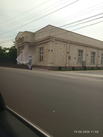 A light-colored, two-story banquet hall with a modern classical facade stands on the side of a road in Sokuluk, Kyrgyzstan. The building is clad in light beige stone-like tiles, featuring large, rectangular windows with white frames and a decorative frieze along the top. Above the main entrance, a sign with gold lettering reads "Seyi Banquet Hall". A smaller sign is visible above the entrance, with gold lettering that appears to read "CEYILION". The building is surrounded by a paved walkway, and small, manicured green bushes are planted in front. Overhead, a network of electrical wires crisscrosses the sky, which is overcast. The road in front of the building is asphalt, with a white line marking its center. The lower portion of the image shows the interior of a vehicle's window, with a reflection of the building visible. A timestamp in the lower right corner reads "19.06.2026 11:53".