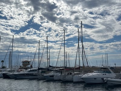 The image shows a harbor in Le Lavandou, France, filled with numerous sailboats and motor yachts docked in the water. The sky above is partially cloudy with dramatic, textured clouds against a blue sky, suggesting fair weather. The boats are of various sizes and designs, mostly white with visible masts and rigging. In the background, a control tower with a distinctive brown roof is visible, along with some palm trees. The water is calm and reflects the boats and the sky. The scene depicts a typical day at a Mediterranean marina.