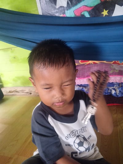 In Pompanua, Indonesia, a young boy is captured in a close-up portrait, his eyes gently closed as if in thought or play. He is seated indoors, the floor beneath him appears to be wooden, reflecting a warm, light brown hue. Behind him, a vibrant, lime-green wall suggests a simple, possibly rustic, interior. Above the wall, a dark blue fabric, possibly a hammock or canopy, stretches across the frame, adding a contrasting color to the scene.

The boy is wearing a t-shirt with a graphic design featuring the word "VICTORY" above a soccer ball. The text "SINCE 1970" is also visible. His right hand is raised, holding a set of keys, which are blurred due to motion. The lighting in the image is soft, with no harsh shadows, indicating ambient indoor light. The overall impression is one of a candid moment, capturing the child in a relaxed and private setting.
