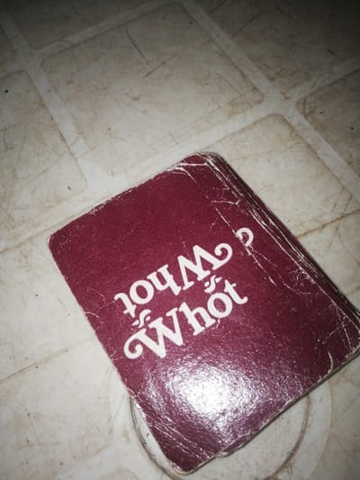 A close-up, angled shot shows a stack of maroon playing cards on a dirty, white, tiled surface. The cards are slightly worn, with visible creases and scuff marks. The top card displays the words "Whot?" in white lettering. The "W" in "Whot?" is a stylized capital letter. The background is a mottled white surface with thin, dark lines and smudges, suggesting a well-used environment. The lighting is uneven, with a bright reflection on the surface of the cards. The context of Ajasse Ipo, Nigeria is not directly observable from the image itself but is provided as external information.