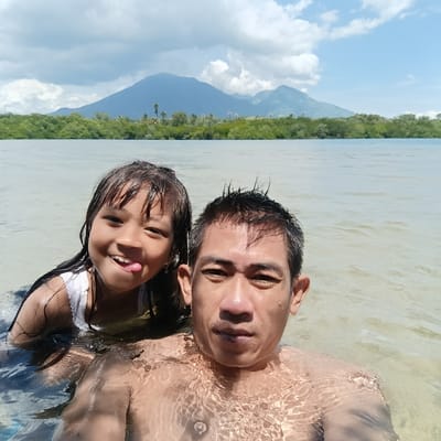 A man and a girl stand chest-deep in clear, shallow water. The girl, to the left, smiles and sticks out her tongue. The man, to the right, looks directly at the camera. Behind them, a coastline of lush green trees stretches across the horizon, with two mountains rising in the background under a cloudy sky. The water's surface creates ripples and patterns on their bodies.