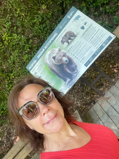 This media file shows a woman taking a selfie while lying down outdoors. She is wearing a red, ribbed top and sunglasses with reflective lenses. A sign about brown bears, titled "MEDVED" - "KHISHNYY VEGETARIANETS" (Bear - Carnivorous Vegetarian), is laid out on the ground in front of her, with images and text in Russian. The sign provides information about bear facts, habitat, and what to do if you encounter a bear. The scene appears to be in a park or natural area, with grass, leaves, and wooden structures visible in the background. The lighting suggests it is daytime. The woman is smiling, and the overall tone is casual and relaxed. The location is indicated as Bolshoy Sochi, Russia.