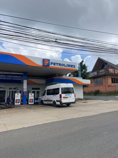 This is a photograph of a Petrolimex gas station located in Cam Ly - Da Lat, Vietnam. The gas station features a blue and orange awning with the Petrolimex logo and name prominently displayed. Multiple gas pumps are visible, along with a white van and a scooter. A person in a blue uniform is standing near the scooter. In the background, a building with a distinctive pitched roof and wooden facade is situated, with a sign that appears to be for a restaurant or cafe. The sky is partly cloudy, and power lines are strung across the upper portion of the image. The scene depicts a typical day at a gas station in this Vietnamese city.