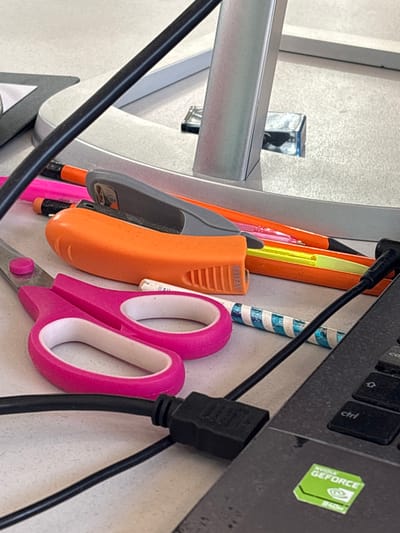 The scene depicts a close-up, elevated view of a desk surface cluttered with various stationery items and parts of electronic devices, located in Tayshet, Russia. The desk surface is a light gray, textured material. In the bottom-left, a pair of pink-handled scissors with silver blades rests open. Immediately above the scissors, an orange plastic stapler with a gray top, bearing the 'AVENUE' brand name on its side, is positioned. Several writing instruments are scattered behind the stapler and scissors: a pink pen/pencil, an orange pen/pencil, and a yellow highlighter. A white pen or marker with blue diagonal stripes is also visible. A thick black cable runs diagonally across the image from the top-left to the bottom-center. To the right, a black laptop is partially visible, showing a portion of its keyboard with 'ctrl' and 'caps lock' keys, and a prominent green 'NVIDIA GEFORCE 840M' sticker. A thinner black cable, connected via a black USB-type connector, plugs into the laptop. In the background, a silver-colored monitor stand or base is visible, and a small, clear, rectangular object reflects light near its base. A dark gray triangular object is partially visible in the upper-left corner. No individuals or specific actions are depicted.