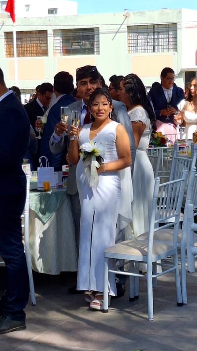 The image captures a wedding celebration occurring outdoors. The location is likely in Cerro Colorado, Peru, as suggested by the architecture and light. The bride, wearing a white dress, holds a bouquet and is holding a glass of champagne. Next to her is the groom, also holding a champagne glass. There are other people present, some in suits and dresses, who are also holding champagne glasses. The wedding table is covered with gifts, decorations and a white table cloth. Numerous white chairs are present. A building with windows can be seen in the background. The scene is bathed in sunlight, casting shadows and highlighting the festive atmosphere.