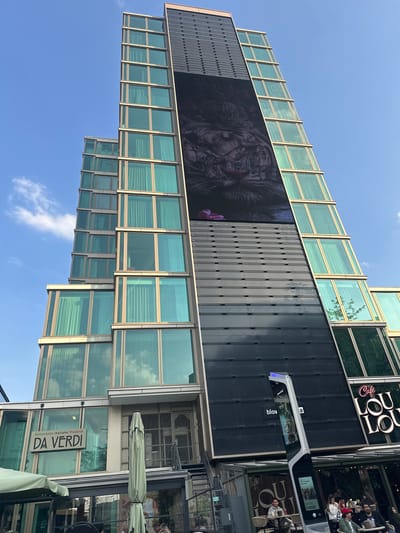 The image depicts a modern architectural structure in Eindhoven, Netherlands, characterized by a series of horizontally stacked blocks, each with a combination of glass-paneled sections and dark, solid sections. The glass panes reflect the sky, which is a clear blue with a few scattered clouds.
One of the dark sections displays a large digital billboard showing a complex, monochromatic image with floral accents. Adjacent to this, another dark section features the signage for "Café Loulou". Further down, a sign for "Da Verdi Ristorante Italiano Pizzeria" is visible, partially obscured by a green umbrella.
In the foreground, there's an outdoor dining area with people seated at tables, suggesting a lively social scene. A "blov" advertisement is displayed on a vehicle next to the outdoor dining area. The architectural design and the presence of these commercial establishments indicate an urban, potentially mixed-use area. The image is taken from a slightly low angle, emphasizing the height of the building.