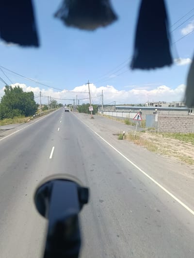 The image shows a view from inside a vehicle, looking out at a road in Jrvej, Armenia. The road is paved and has white lane markings. There are trees and vegetation on the left side of the road, and buildings and a fence on the right. The sky is blue with some clouds, suggesting it is daytime and the weather is fair.

There are a few objects visible: a car driving in the distance, and a traffic sign on the right side of the road. The traffic sign is a triangular warning sign with a symbol indicating a crossroads or T-junction. There is also text on a sign in Georgian script on the right side of the road. The foreground of the image is dominated by the blurred outline of the vehicle's interior, possibly a windshield mount for a device.

The overall scene appears to be a drive along a semi-rural road. There are power lines visible overhead, indicating infrastructure in the area. The presence of buildings under construction on the right suggests development. The time of day appears to be late morning or early afternoon, given the bright sunlight. There are no people clearly visible in the image.