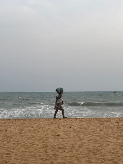 The image displays a beach scene with an overcast sky, the ocean, and a sandy shore. The photograph appears to have been taken in a portrait orientation but is currently rotated 90 degrees clockwise, placing the sky on the left, the ocean in the middle, and the sand on the right.
When viewed with the correct orientation:
The top third of the image is dominated by a light grey, overcast sky, suggesting either a cloudy day or a time when the sun is not directly visible. There are no distinct cloud formations or blue patches, indicating a uniformly muted light.
Below the sky lies the expanse of the ocean. The water is a dark, somewhat murky blue-green color, with white foamy waves breaking closer to the shore. Further out, the water appears calmer with subtle ripples.
The bottom portion of the image is a sandy beach, characterized by its light brown color and visible textures from the water and wind.
A single person, appearing to be a child or young individual, is positioned centrally in the frame, standing on the wet sand at the water's edge. The person is dressed in a light-colored, possibly striped, short-sleeved top and dark patterned shorts or a skirt. A colorful cloth, featuring shades of blue and green, is wrapped around their head like a turban or head covering. Their arms are bent at the elbows, with hands slightly raised, giving an impression of being in a state of balance or momentary pause. The person is facing slightly towards the left side of the frame, looking out at the ocean.
The overall atmosphere is calm and serene, with the soft, diffused lighting from the overcast sky. No specific text or identifiable landmarks are visible in the image.