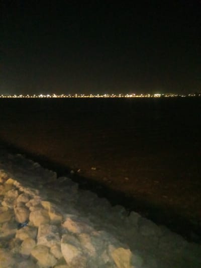 This nighttime photograph presents a coastal view, likely taken from the shore of Al Khobar, Saudi Arabia. In the immediate foreground, a rugged bank of large, light-colored rocks occupies the lower portion of the frame, suggesting a sea wall or natural rocky coastline. A small blue object, possibly a piece of litter, is visible nestled among these rocks in the bottom left corner.
Beyond the rocky bank, a wide expanse of dark water fills the middle ground, indicating a large body of water such as the Arabian Gulf. The water's surface is predominantly dark, reflecting minimal light.
Stretching across the horizon in the distance is a distinct, linear array of bright lights, illuminating what appears to be a distant cityscape or a developed coastline. These lights are predominantly yellow and white, forming a vibrant streak against the dark background. The sky above is a deep, unlit black, suggesting a clear night with no visible moon or cloud cover.
No people or specific activities are visible in the image. The scene conveys a quiet, expansive view of an urbanized waterfront at night, characterized by the contrast between the dark natural elements and the distant, glowing artificial lights. There is no visible text.