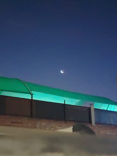 A crescent moon hangs in a dark blue sky above a structure with a green awning. Below the awning is a wall made of dark-colored panels with vertical support beams, resting on a brick base. In the foreground, a concrete surface is visible. There is a small point of light in the top left. No humans visible. The location is not identifiable as Trujillo, Peru based on visual information.