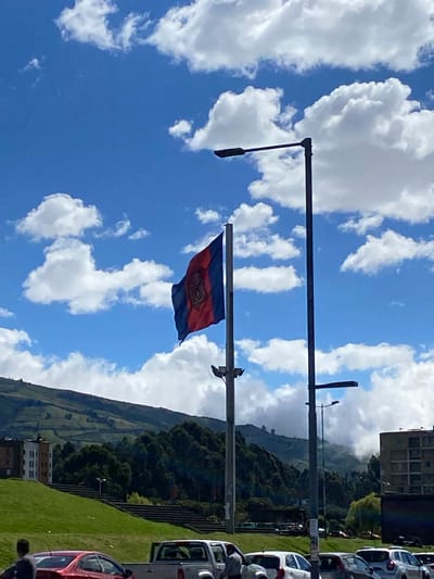 The image shows a flag with a blue and red horizontal stripe design flying from a pole. The sky is blue with scattered white clouds. In the background, a lush green hillside rises, possibly a mountain. There are cars parked on the side of the road. The location is likely Quito, Ecuador, based on the flag and landscape.