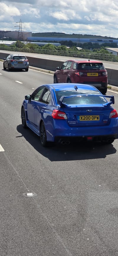 A blue Subaru WRX sports sedan is captured from a rear-quarter view on a highway, likely the M1 northbound, as indicated by the presence of "PRETTYLITTLETHING.COM" billboards in the background, a known distribution center for the company located near Sheffield, United Kingdom. The Subaru, displaying the license plate "X200 OPW", is in the middle lane, accompanied by a red Land Rover Discovery SUV in the right lane with license plate "YJ18 WRV", and a silver hatchback in the left lane, partially obscured. The sky is partly cloudy, suggesting daytime conditions. The road surface is asphalt, marked with white lines and reflective road studs. The context suggests a typical highway driving scenario.