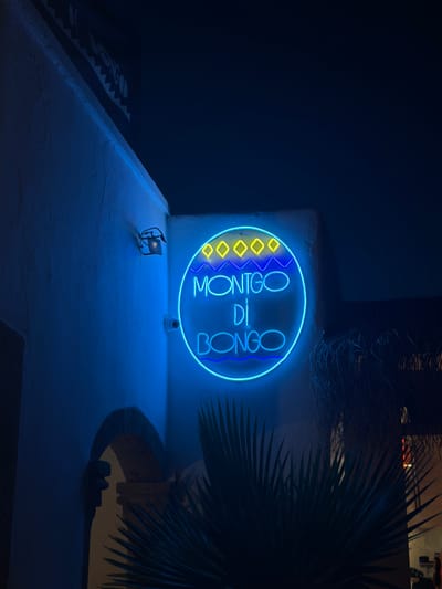 The image shows a bright blue neon sign that reads "MONTGO DI BONGO". The sign is circular with yellow diamond shapes on the left side and a blue wavy line on the right. It is mounted on a white, textured wall in an outdoor setting at night. In the foreground, a white concrete surface is visible, with a small black metal object resembling a miniature chair or sculpture. To the right, lush green palm fronds are visible, obscuring part of the background. Behind the neon sign, a dimly lit architectural structure with arched openings and interior lighting can be seen, suggesting a building or venue. The overall ambiance is nocturnal and atmospheric, with the neon light casting a vibrant blue glow. The location context of Xàbia/Jávea, Spain, suggests a coastal or Mediterranean setting. The image appears to be taken from an elevated perspective, looking down onto the illuminated sign and the surrounding area. There are no people visible in the frame.