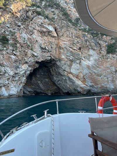 The image captures a close-up view from a boat navigating near a steep, rocky cliff face with a dark cave opening. The scene is set in Skopelos, Greece, a location known for its dramatic coastlines. The rocky cliff dominates the left side of the frame, showing textured layers of stone in shades of brown, grey, and white, with sparse vegetation clinging to its surface. The water, a deep blue-green, is visible on the right, lapping against the hull of a white boat.
Part of the boat is in the foreground, showing a white deck, metal railings, and cleats. A bright orange life preserver with white stripes is secured to the railing. A canvas sunshade is also visible. The angle of the shot suggests the boat is moving slowly alongside the cliff, likely exploring the coastline or approaching the cave.
The lighting indicates it is daytime, with sunlight casting shadows on the rocks, highlighting their rugged contours. The water appears calm, suggesting fair weather. The overall impression is one of natural beauty and exploration, with the imposing cliff and the dark cave hinting at the geological history of the area. A small yellow warning sticker with black text is visible on the boat's hull, with the words "CAUTION" and "SLIPPERY WHEN WET" legible.