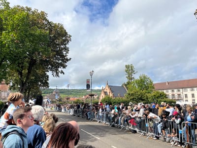 An outdoor crowd is gathered on a street in Molsheim, France, observing an event. The scene is set on what appears to be Rue de la Commanderie, with historical buildings in the background. On the left, partially obscured by large green trees, is a historical building with a dark roof and a distinctive small, ornate spire, identified as the Chartreuse de Molsheim. To its right, a large, multi-story building with a light-colored facade and reddish-brown tiled roof, characteristic of the Commanderie Saint-Jean, is visible. Distant hills covered with vineyards are visible behind these structures.

Approximately one hundred spectators of various ages, including adults and children, are visible along the right side of the frame and extending down the street. They are standing behind grey metal crowd control barriers that line both sides of the asphalt road. Many individuals are leaning forward against the barriers, looking down the street in anticipation. One child, wearing a multicolored shirt, is seated on an adult's shoulders in the foreground, facing away from the camera. People are dressed in casual clothing.

On the right side of the street, a dark street lamp post features a black banner with a white Bugatti logo, indicating the event's association with the luxury car manufacturer. Further down the street, a blue archway structure is visible, potentially marking a start or finish line. The sky is partially cloudy with patches of blue.
