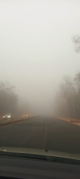The image captures a two-lane asphalt road in Ait Naamane, Morocco, enveloped by dense fog, severely limiting visibility. The perspective is from inside a vehicle, with a portion of the dashboard visible at the bottom of the frame. A faded white line marks the center of the road. On the left shoulder, a light-colored sedan is visible, facing away from the viewer, with its taillights illuminated. Further ahead on the road, two pairs of amber headlights are faintly discernible through the fog, suggesting oncoming traffic. On both sides of the road, the silhouettes of bare-branched trees are barely visible, partially obscured by the atmospheric conditions. The sky is uniformly white due to the fog, which dominates the entire environment.