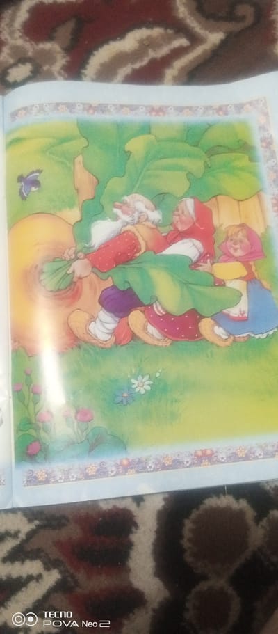 The image is a colorful illustration of three people pulling on a giant turnip. The scene takes place in a grassy field with a blue sky and some flowers. The characters appear to be a grandfather, grandmother, and a granddaughter, dressed in traditional clothing. The grandfather is an elderly man with a white beard and is wearing a red polka-dot shirt and purple pants. The grandmother has a red headscarf and a green dress. The granddaughter is wearing a pink headscarf and a blue dress. They are all pulling on the turnip with great effort. The turnip is enormous and appears to be the focal point of the illustration. A small blue bird is flying in the background. The illustration is bordered by a decorative pattern of flowers. The image is likely from a children's book or story. The location is not discernible from the image.