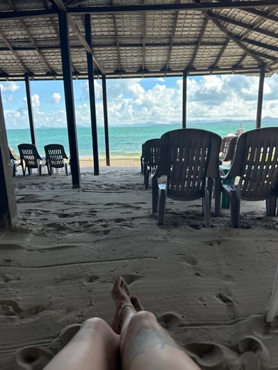 The image depicts a scenic beach setting in Ban Khlong Hia, Thailand. In the foreground, the sandy beach is marked with footprints, suggesting recent activity. A person's legs and feet are visible on the right side of the frame, adorned with a bracelet on one ankle. Beyond the sand, several rows of empty, dark plastic chairs are arranged under a rustic, open-air wooden structure with a thatched roof. These chairs face the ocean, hinting at a setup for relaxation or dining with a view.

The sea is a vibrant turquoise, stretching towards a distant shoreline with hazy, mountainous terrain. The sky above is a mix of bright blue with scattered white clouds, indicating a pleasant, possibly sunny day. The overall impression is one of tranquility and a relaxed atmosphere, characteristic of a beachside establishment. There is no visible text in the image. The time of day appears to be daytime, given the lighting and the clear sky. The location cues, such as the tropical vegetation visible in the distance and the architecture of the shelter, strongly suggest a Southeast Asian coastal environment.