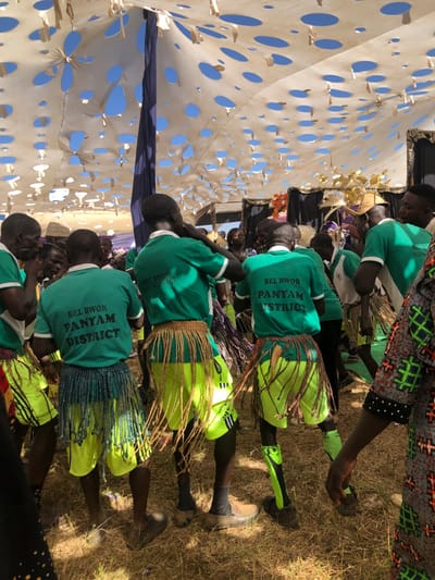 Under a tent with a white fabric roof spotted with circular cutouts, a line of people wearing green shirts and yellow skirts with fringed edges stand. The shirts bear the words "BEL BWOR PANYAM DISTRICT". The people are primarily viewed from behind, with glimpses of their dark hair. The ground appears dry and grassy.