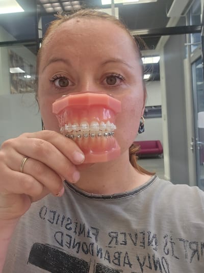 A light-skinned individual with reddish-brown hair, wearing a light grey t-shirt with dark text, holds a dental model with braces in front of their face. The background shows a room with reflective surfaces, including a window with square divisions. Behind the individual, a partially visible pink object is noted. The location is identified as Mouscron, Belgium, based on metadata that is not present in the image itself.