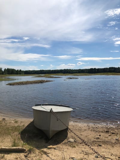 A small, white boat is pulled up onto a sandy shore next to a body of calm water. The boat is secured with a metal chain. In the background, a line of trees forms the horizon, and a bright blue sky with scattered white clouds is visible. The scene appears to be a serene outdoor location, possibly along the banks of the Svir River in Svirstroy, Russia, given the characteristic landscape and vegetation. The overall lighting suggests it is daytime, likely mid-morning or afternoon. There are no people visible in the image.