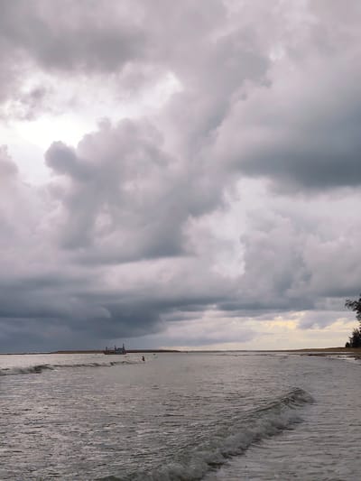 The image depicts a beach scene in Sakhu, Thailand, under a dramatic, overcast sky filled with heavy, grey clouds. The sea is calm, with gentle waves lapping at the shore. In the distance, a small fishing boat is visible, and a solitary figure can be seen in the water near the boat, possibly swimming or wading. The coastline is lined with low-lying vegetation and trees on the right side of the frame. The overall atmosphere is one of impending rain or a storm, with muted light and a somber color palette.