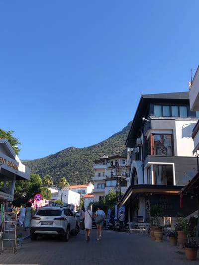 This image depicts a street scene in Kaş, Turkey, under a clear blue sky. A white Toyota SUV, license plate 07 Z 6700, is parked on the right side of the street. Several people are walking on the sidewalk. A white building with a dark, modern facade and a balcony with a metal railing is prominent on the right side of the image. To the left, a building with a white awning has text that appears to read "ENT BÖREK". In the background, a densely wooded hillside rises towards the sky. Palm trees are visible, along with other buildings in varying architectural styles. The overall atmosphere suggests a sunny day in a coastal town.
