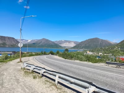 This image captures a scenic view on a sunny day, likely near Kirovsk, Russia, given the mountainous terrain and the presence of a body of water. A paved road curves through the landscape, bordered by a metal guardrail. On the left side of the road, a light pole with wires is visible, along with a circular sign attached to a wooden post. Beyond the road, a wide, blue body of water stretches towards distant mountains. These mountains are characterized by their rugged slopes, some of which are dusted with snow, indicating a cool climate. Scattered in the foothills and along the shoreline are buildings, suggesting a small settlement or town. Lush green vegetation, including trees and bushes, thrives around the water's edge and on the lower slopes of the mountains. The sky is a clear, vibrant blue with a single, wispy cloud. The overall impression is one of a remote, natural landscape with signs of human habitation.