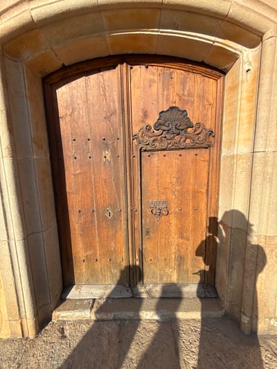 The image shows a close-up, frontal view of a weathered wooden double door set within a stone archway. The doors are made of vertical wooden planks, studded with small, dark metal fasteners. The right door has an ornate black metal decorative element at the top and a metal handle and keyhole below. The stone archway and surrounding wall are a pale, textured stone, with a curved arch at the top. Sunlight casts strong shadows across the scene, including the shadows of two people, one of whom is holding a phone and appears to be taking a photo. The ground in front of the door is paved with rough stone. The context of Tossa de Mar, Spain, suggests this is likely an exterior architectural feature of an older building in the town, possibly within the Vila Vella, the walled medieval town.