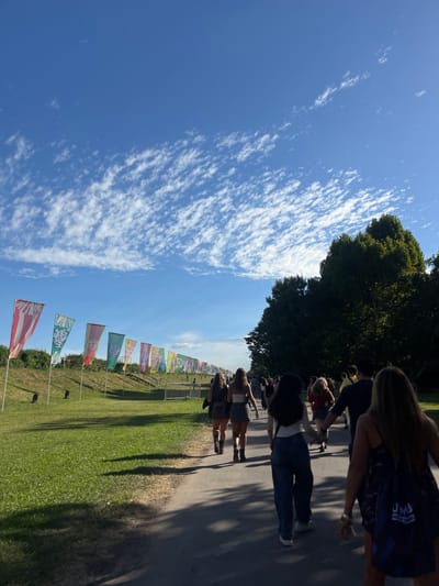 The scene depicts an outdoor event pathway at the Hipódromo de San Isidro in San Isidro, Argentina. A bright blue sky with scattered white altocumulus clouds is visible overhead. Direct sunlight casts long shadows to the viewer's right, suggesting late afternoon. Approximately 15-20 young adults are walking away from the camera on a paved path that recedes into the distance. Individuals are dressed in casual, warm-weather attire, including shorts, skirts, and t-shirts; one person wears jeans. Some walk in pairs or small groups; a couple is observed holding hands in the mid-ground. To the left of the path, a grassy embankment rises, lined with approximately 15 brightly colored, vertical rectangular flags mounted on poles. The second flag from the left partially displays the text "Lollapalooza," indicating a music festival. Metal crowd control barriers are visible further along the path and behind the flags. Two small, black, cylindrical objects are positioned on the grass in the left foreground. A dense line of tall, green deciduous trees borders the right side of the path.
