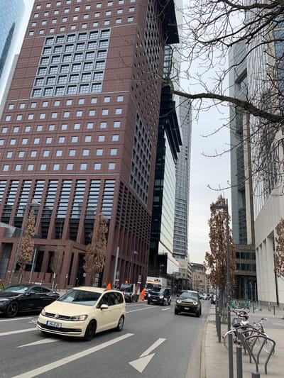 The image shows a street scene in the city of Frankfurt am Main, Germany. The location is the intersection of Neue Mainzer Straße and Taunusanlage (latitude: 50.111, longitude: 8.672). The image captures a light cream colored minivan traveling to the left and a dark colored SUV traveling to the right on the street. There is a taller modern office building on the left of the image and a taller modern office building on the right of the image. The buildings appear to be constructed of red brick with numerous windows. There are a series of trees with no leaves on them along the curb of the street and several bicycles parked on the sidewalk.