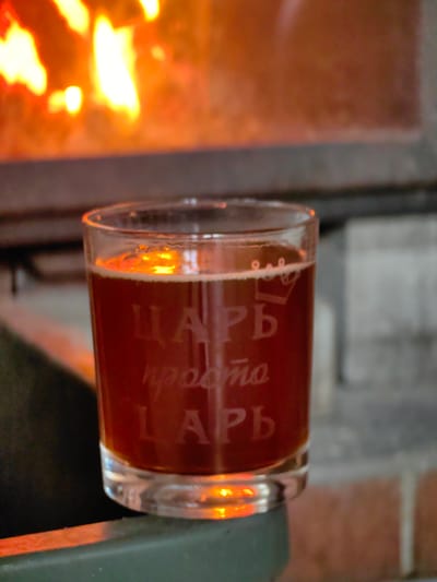 A close-up, eye-level shot focuses on a glass of dark amber liquid, likely beer, with a frothy head. The glass is positioned in the foreground, resting on a dark green surface that appears to be part of a fireplace or stove. The glass is etched with text that reads "ЦАРЬ просто ЛАРЬ". Above the glass, a warm, blurry glow emanates from a fire, with bright orange and yellow flames visible, suggesting a cozy indoor setting. The background consists of a rough, brick-like texture in muted tones, further supporting the idea of a fireplace. The lighting is warm and ambient, primarily driven by the fire's illumination. The image is taken in the city of Фаніпальскі сельскі Савет, Belarus.